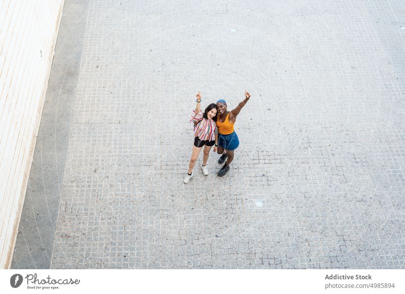 Stilvolles multiethnisches Paar von lesbischen Frauen in der Stadt cool urban lgbt Lächeln erhobener Arm heiter positiv gleichgeschlechtlich Freundin Straße