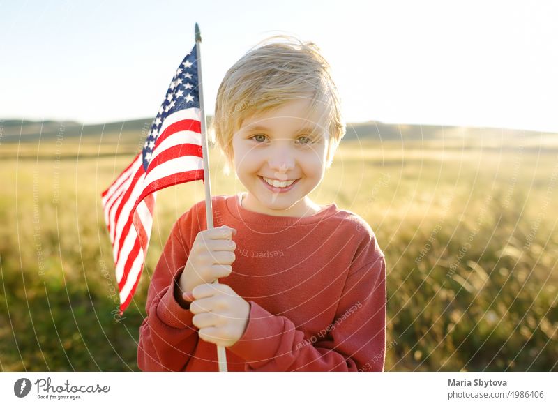 Netter kleiner Junge feiert den 4. Juli, den Unabhängigkeitstag der USA bei sonnigem Sommer Sonnenuntergang. Glückliches Kind läuft und springt mit amerikanischer Flagge Symbol der Vereinigten Staaten über Weizenfeld.