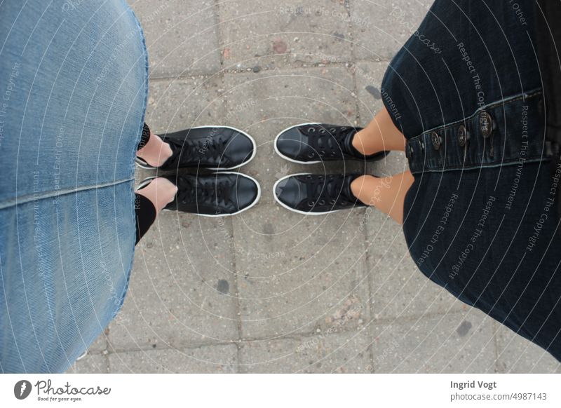 Zwei Paar schwarze Turnschuhe und zwei Jeansröcke; zwei Frauen stehen sich gegenüber, Vogelperspektive Schwarze Turnschuhe Füße Schuhe von oben Steinplatten