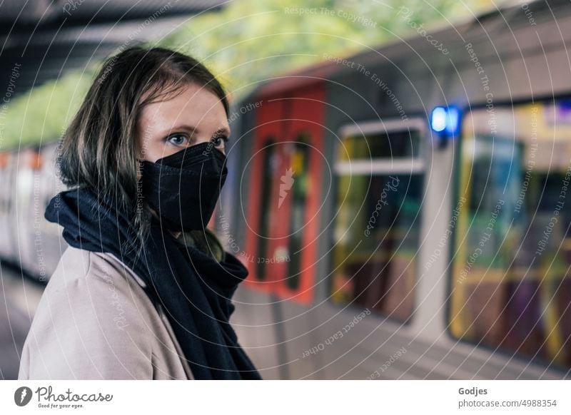 Junge Frau mit Mundschutz steht auf einem Bahnsteig und wartet auf den ankommenden Vorortzug | Corona Gedanken Korona Bahnhof SBahn Corona-Virus unterirdisch