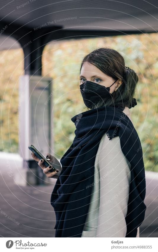 Junge Frau steht auf einem Bahnsteig mit Smartphone in der Hand und trägt einen Mundschutz | Corona-Gedanken Bahnhof SBahn Eisenbahn Außenaufnahme Farbfoto