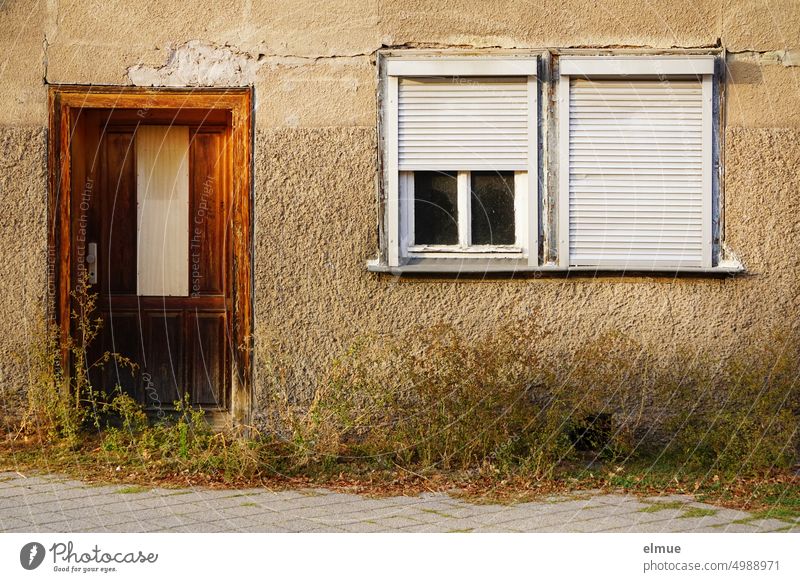 marodes Wohnhaus mit alter Holztür, zwei Fenstern mit halb bzw. ganz heruntergelassener Jalousie und zahlreichem Unkraut davor verlassen kaputt Wandriss Fußweg