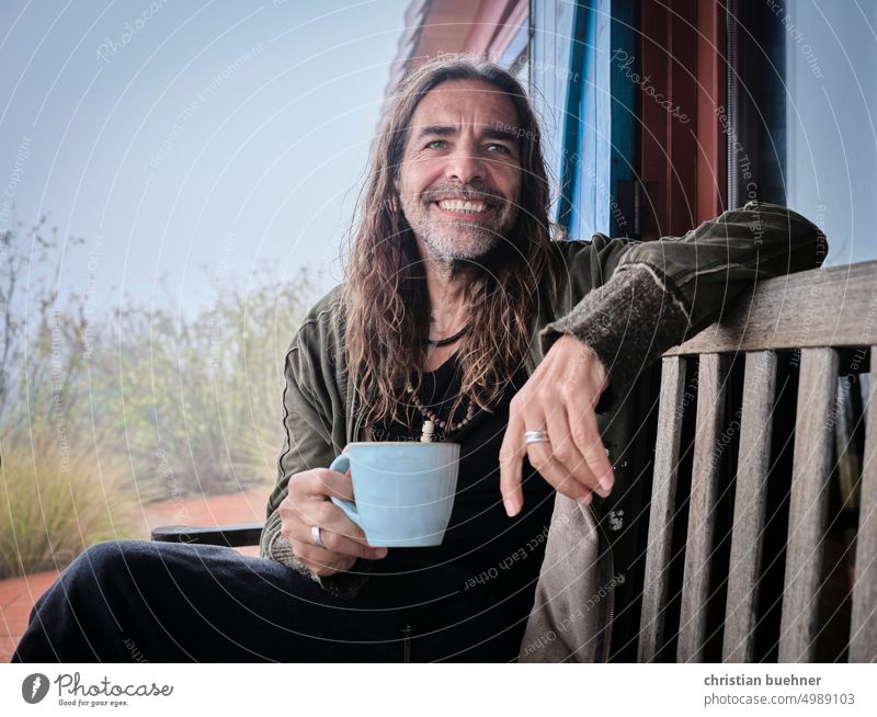 maenner portrait mit tasse mann lange haare langhaarig hippie teetasse kaffetasse kaffepause freundlich lachen fröhlich lachend bewölkt regen natur holzbank