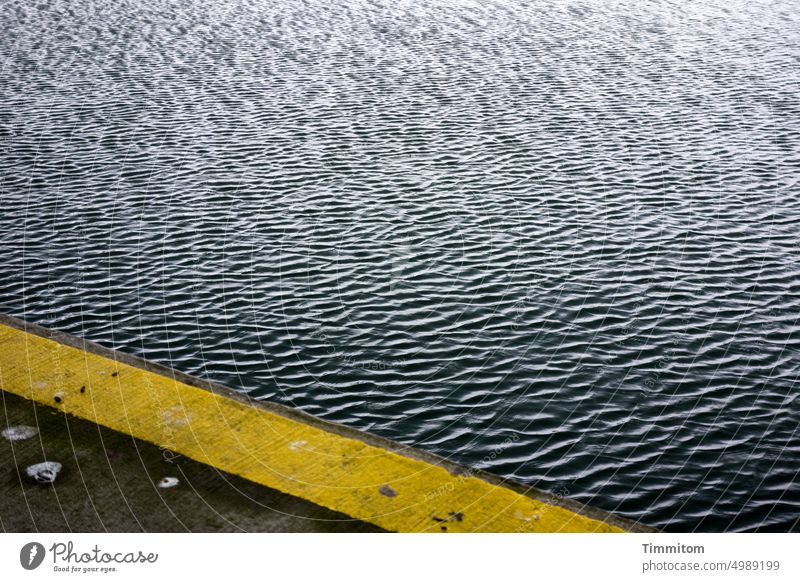 Kleine Wellen am Hafenrand Betonboden Abgrenzung Markierung gelb Wasser dunkel Licht Schatten Menschenleer Dänemark Vogelkot