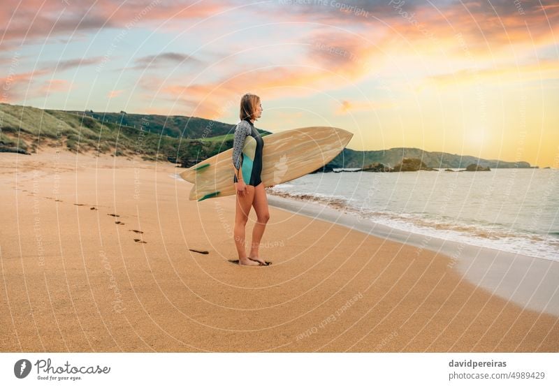 Surferin mit Neoprenanzug und Surfbrett am Strand mit Blick auf das Meer jung Frau MEER Rücken Sand Fußspuren posierend tragen Menschen 20s im Freien Sport