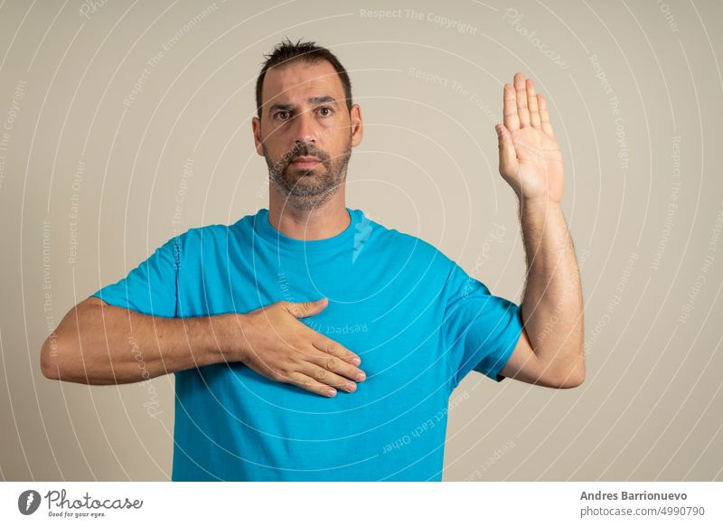 Bärtiger hispanischer Mann in den 40ern mit blauem T-Shirt auf beigem Hintergrund, der mit der Hand auf der Brust und offener Handfläche schwört, den Treueeid zu leisten.