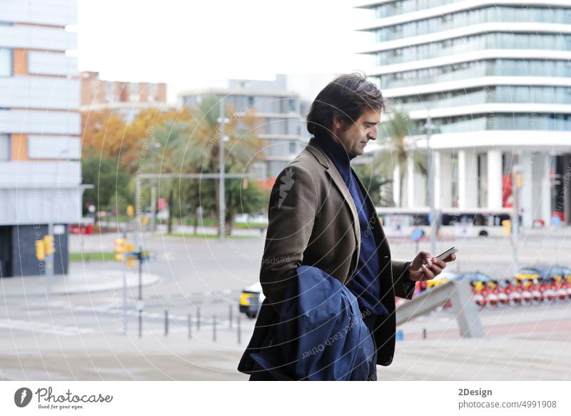 Selbstbewusster Mann, der auf sein Handy schaut, während er auf einer Straße spazieren geht, Mann im Anzug, der auf einer Straßenmauer mit Kopierraum läuft