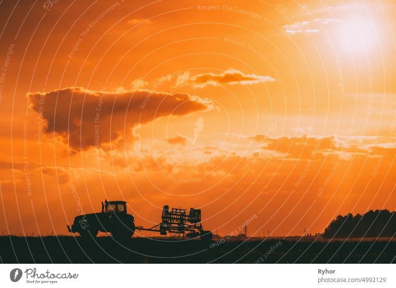 Traktorfahrten auf der Landstraße. Beginn der landwirtschaftlichen Frühjahrssaison. Cultivator Pulled By A Tractor In Rural Field Landscape Under Sunny Summer Sunset Sunrise Sky. Backlit Dramatische Beleuchtung