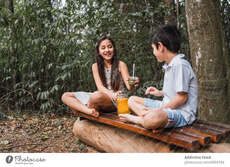 Lächelndes Mädchen übergibt Kumpel an Bruder auf Bank gegen Bäume Pass Gefährte trinken Getränk Erfrischung interagieren Beine gekreuzt heiter Kräuterbuch Tee