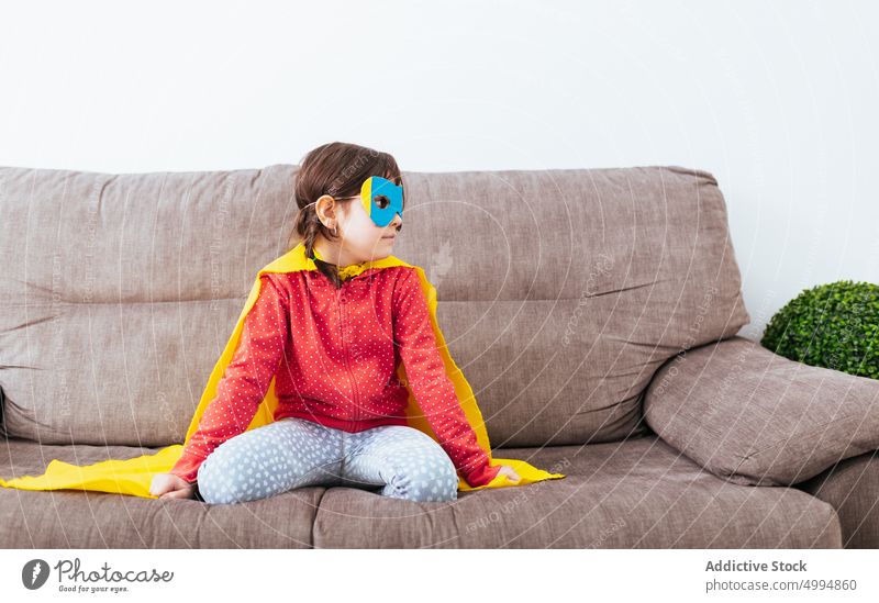 Mädchen mit Maske und Umhang, das vorgibt, ein Superheld zu sein Kap Mundschutz Tracht so tun, als ob selbstbewusst Kindheit Raum Sofa Licht heimwärts weiß Wand