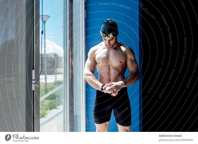 Muskulöser Schwimmer mit nacktem Oberkörper in der Nähe des Fensters, der auf seine Smartwatch schaut Athlet nackter Torso Sixpack Bauchmuskeln verträumt Macho