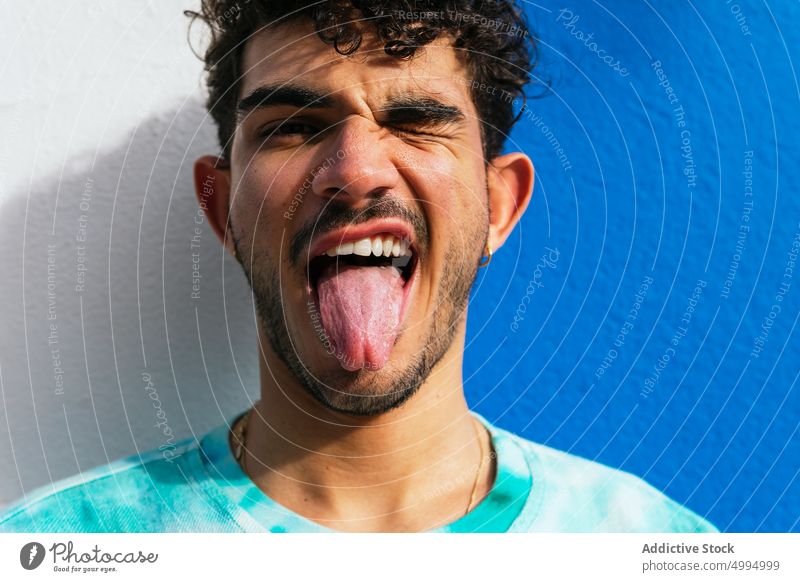 Lustiger hispanischer Mann zeigt Zunge Zunge zeigen spielerisch Zwinkern Grimasse lustig Wand urban Straße Porträt männlich ausspannen Gesicht machen ethnisch