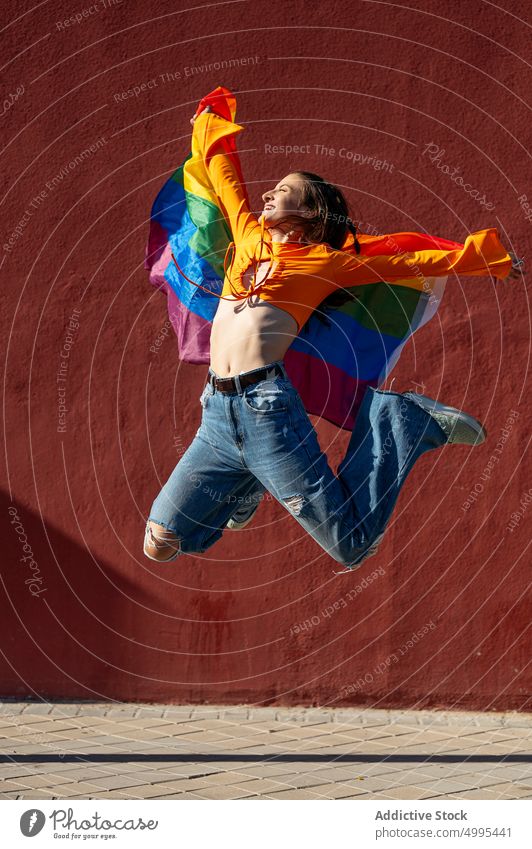 Junge Lesbe springt mit LGBT-Flagge Frau lesbisch springen lgbt Fahne Stolz Straße Glück Wand Freiheit jung farbenfroh Lächeln Arme hochgezogen Regenbogen urban