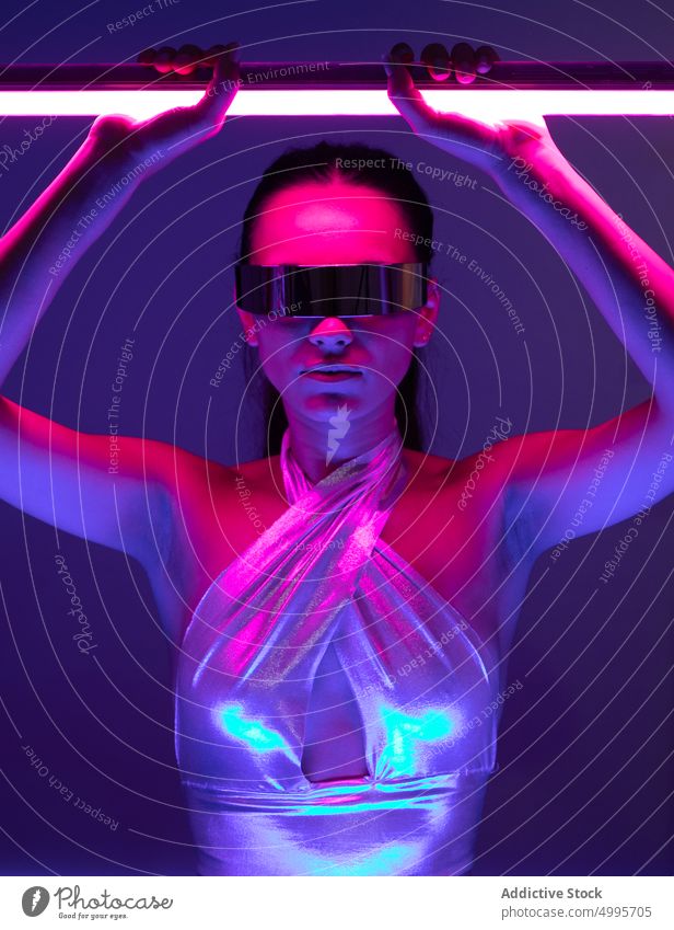 Futuristische junge Frau mit trendiger verspiegelter Sonnenbrille und Neonstäben im Studio Virtuelle Realität futuristisch selbstsicher Stil selbstbewusst
