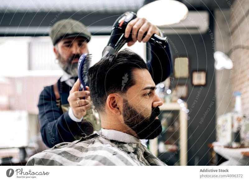 Ethnischer Friseur mittleren Alters trocknet das Haar eines Kunden in einem Friseursalon Männer Klient ernst Haartrockner Haarschnitt Kamm Salon Konzentration