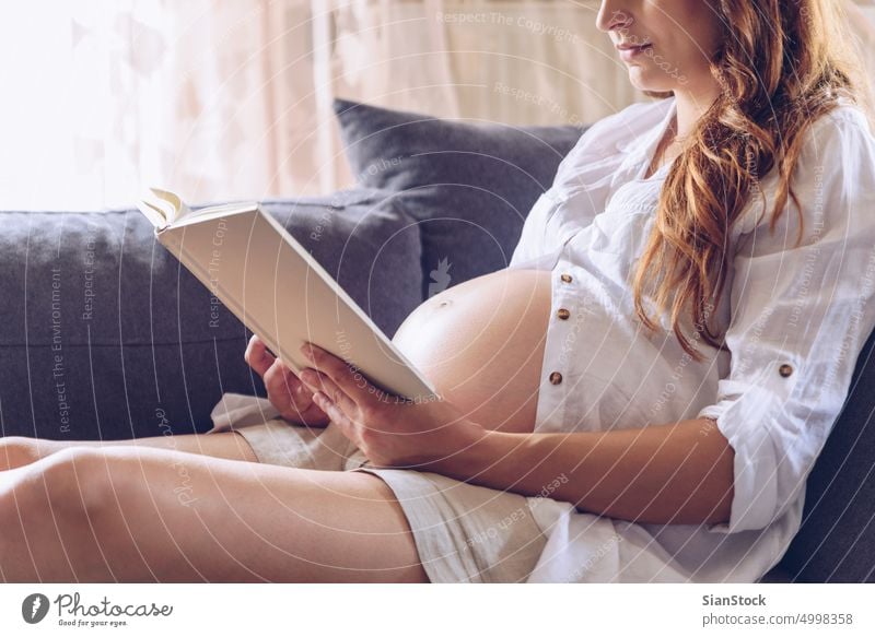 Schwangere Frau, die in ihrem Haus ein Buch liest. weiß Sofa Wohnzimmer Sitzen gemütlich Schlafzimmer Morgen Sonntag Lügen heimwärts Schwangerschaft schwanger