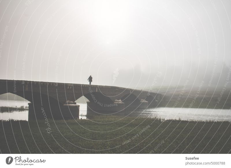 Ein einsamer Mensch im Nebel geht über eine Brücke. Im Hintergrund scheint die Sonne durch den Nebel. Landschaft Wasser allein Spaziergänger alt Mauer Bauwerk