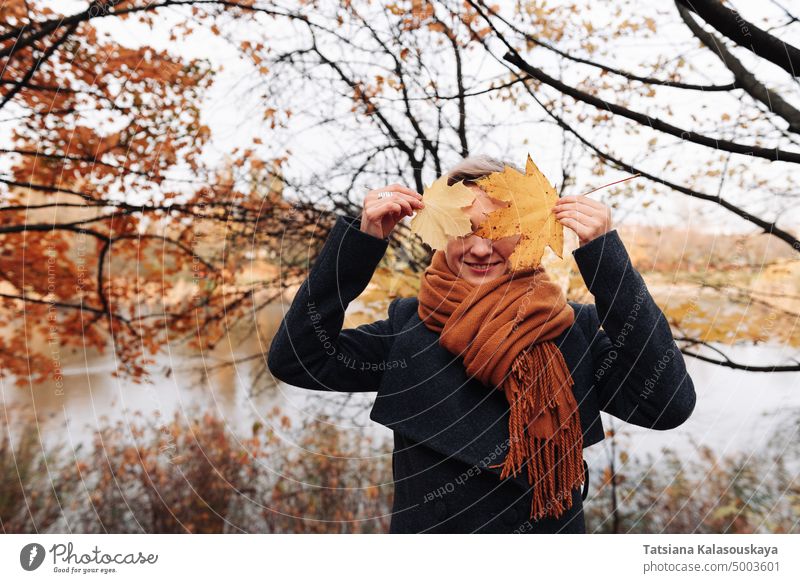 Eine Frau in Mantel und Schal in einem herbstlichen Park bedeckt ihr Gesicht mit Ahornblättern versteckend verdunkelt deckend vor Ahorne Blätter