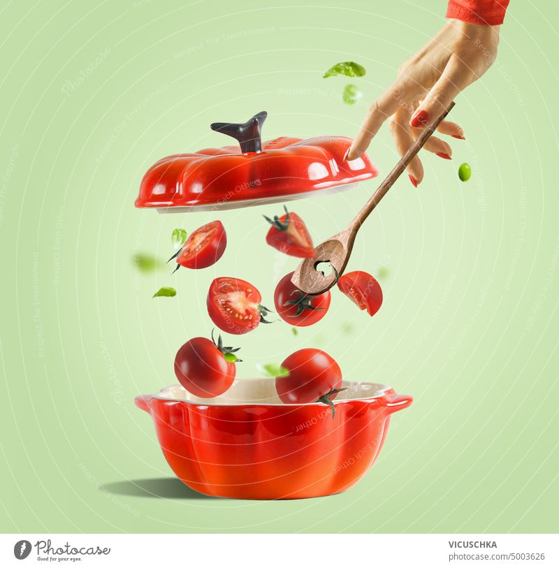 Zubereitung von Tomatensauce oder Tomatensuppe. Roter Kochtopf mit offenem Deckel und fallenden Tomaten und Frauenhand mit Löffel auf hellgrünem Hintergrund. Lebensmittel Levitation.