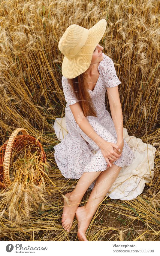Junge barfüßige Frau in weißem Kleid und Hut sitzt im Getreidefeld Sonne Feld Natur Sommer Barfuß Korb Ansicht Weizen Roggen Behaarung blond Weide ländlich