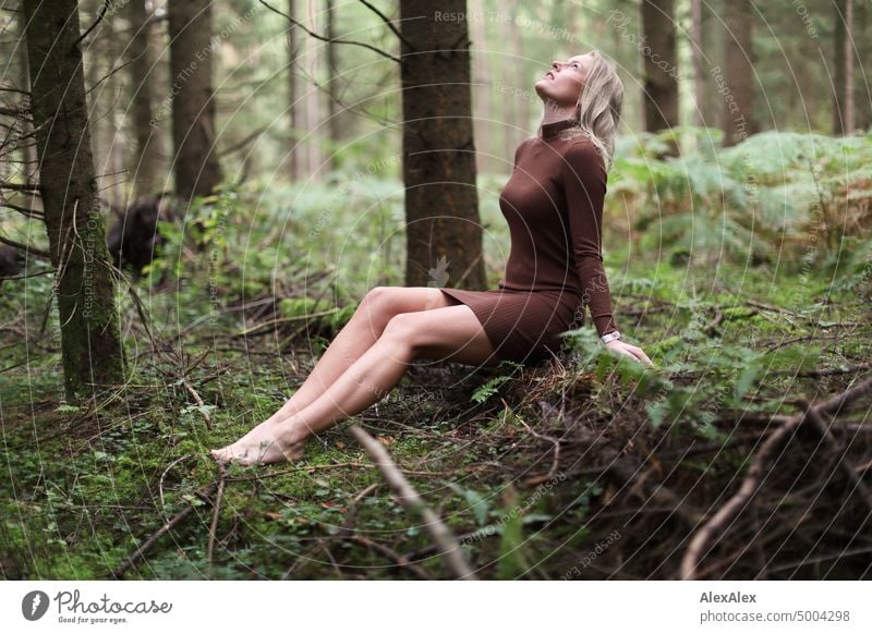 Junge, blonde Frau sitzt barfuß auf einem umgefallenen Baumstamm im Wald und schaut in den Himmel junge Frau feminin schön glücklich Jugendliche Portrait