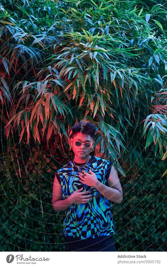 Stilvolle nicht-binäre Person unter Neonlicht Frau Transgender nicht binär neonfarbig Abend Buchse leuchten Garten Vorschein Sonnenbrille Strauch feminin