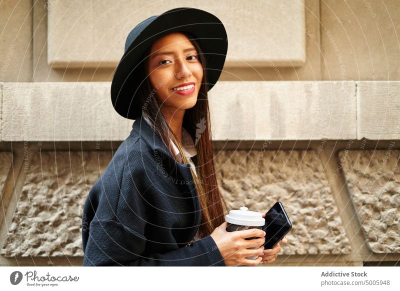Fröhliche hispanische Frau mit Kaffee zum Mitnehmen und Smartphone Stil trendy Mode Imbissbude Gebäude Straße Großstadt Funktelefon Telefon Lächeln Outfit