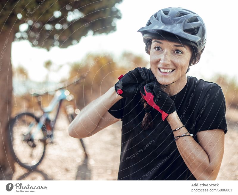 Lächelnde Frau knöpft Helm und Fahrrad in der Nähe eines Baumes zu Schutzhelm knöpfend Sport extrem Aktivität Handschuhe Sicherheit Wald heiter jung Dame