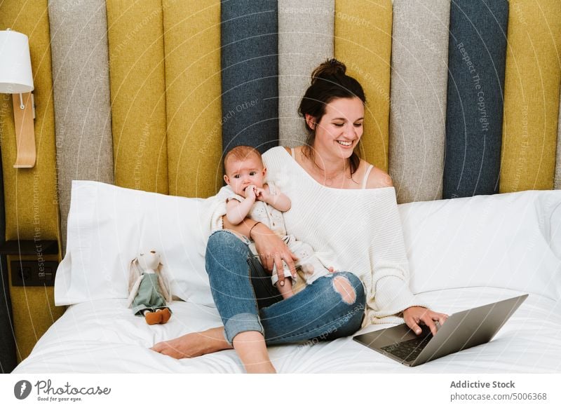 Fröhliche Mutter, die den Computer mit Interesse benutzt, während sie mit ihrem Kleinkind zu Hause chillt Laptop Baby Eltern Kind heimwärts heiter benutzend