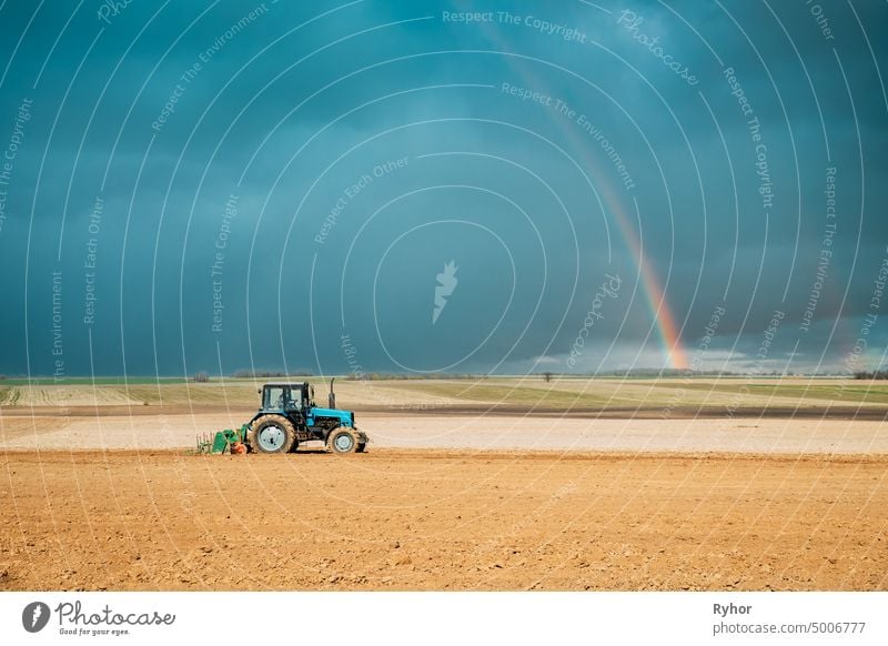 Traktor Pflügen Feld im Frühjahr Saison. Beginn der landwirtschaftlichen Frühjahrssaison. Cultivator Pulled By A Tractor In Countryside Rural Field Landscape Under Spring Blue Sky With Rainbow After Rain