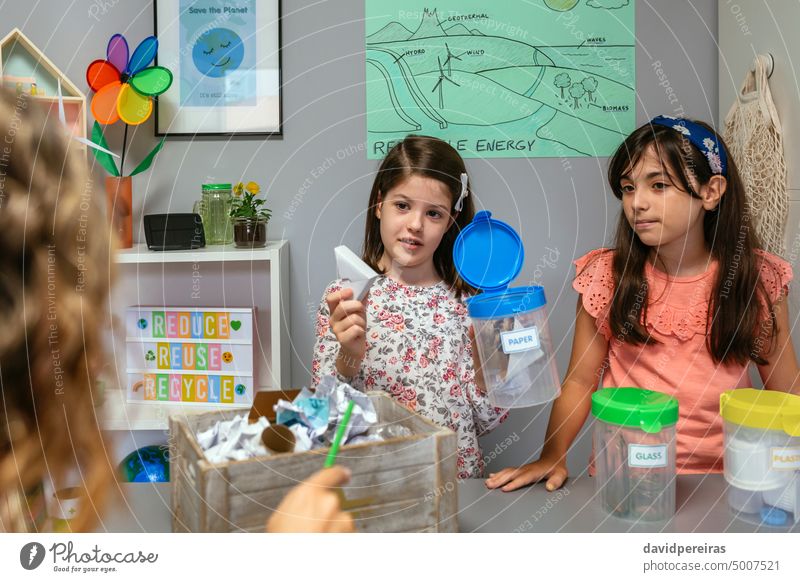 Ein Schüler erklärt, wie man Pappe recycelt, während er die Mülltonne sortiert Mädchen zu erklären. wiederverwerten Lehrer Karton Papier Ökologie Beteiligung