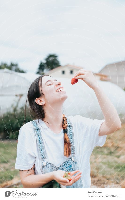 Junge Frau mit frischen Erdbeeren im Gewächshaus, isst und spielt mit ihnen, zeigt die frischen Früchte in die Kamera. Ländliche Arbeit und Ressourcen Konzept. hart arbeitende junge Frau