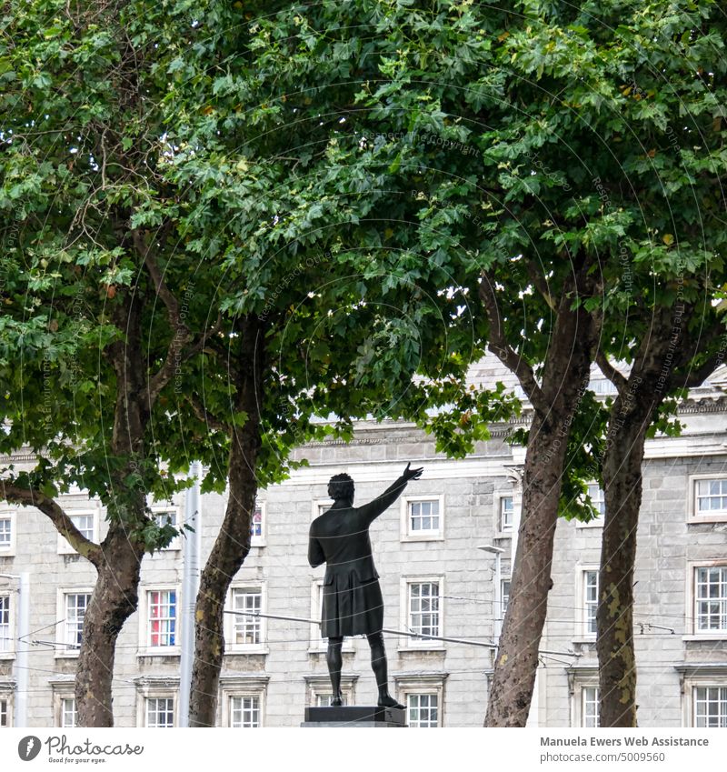 Eine Statue unter Bäumen mit erhobener Hand vor einem großen Gebäude in Dublin statue redner erhobene hand gebäude bäume irland dichter dichten vortragen