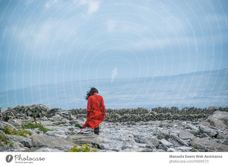 Eine Frau mit Kopfhörern geht an einem Steinstrand spazieren, ihr rotes Kleid wird vom Winde verweht vom winde verweht kleid frau gehen musik hören kopfhörer