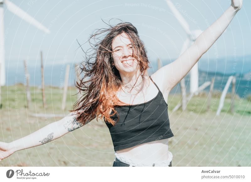 Frontalporträt glückliche junge Frau, die die Freiheit genießt, die Arme in den Himmel hebt und in die Kamera lächelt, umgeben von elektrischen Windrädern. Blasen Haare und entspannen, saubere Erde Zukunft Konzept