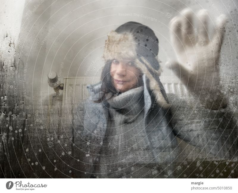 Eine frierende Frau sitzt mit dicker Winterkleidung vor einer Heizung und schaut durch das beschlagene Fenster heizen Energie Energie sparen Fensterscheibe