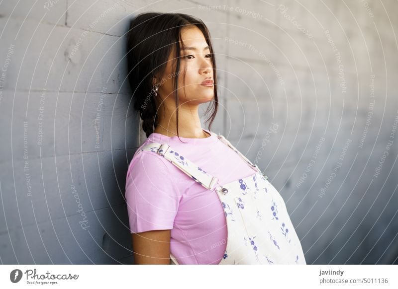 Junge Chinesin mit verlorenem Blick und ernster Miene, die an der Wand eines modernen Bürogebäudes lehnt. asiatisch Chinesisch Frau Japanisch Mode Person