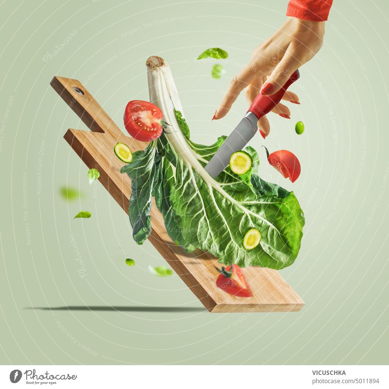 Frauenhand mit Messer, fliegende Salatzutaten und Schneidebrett auf hellgrünem Hintergrund. Kreative Lebensmittel Levitation. Gesundes Essen und Diäten Hand