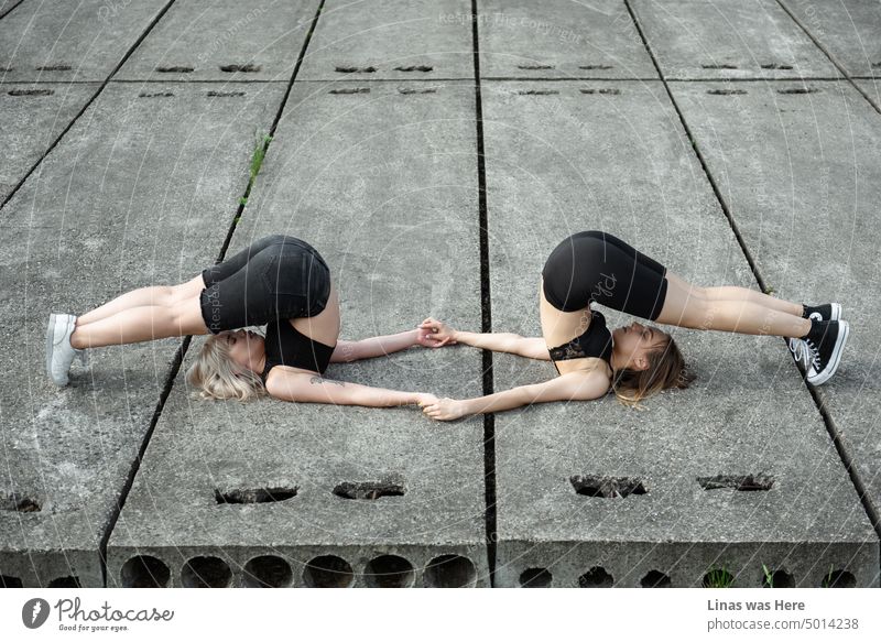 Hübsche Mädchen halten sich an den Händen und machen eine Art Yoga. Auf einem Betonboden. Mit geschlossenen Augen. Diese wunderbaren Freundinnen sind verbunden und konzentriert.