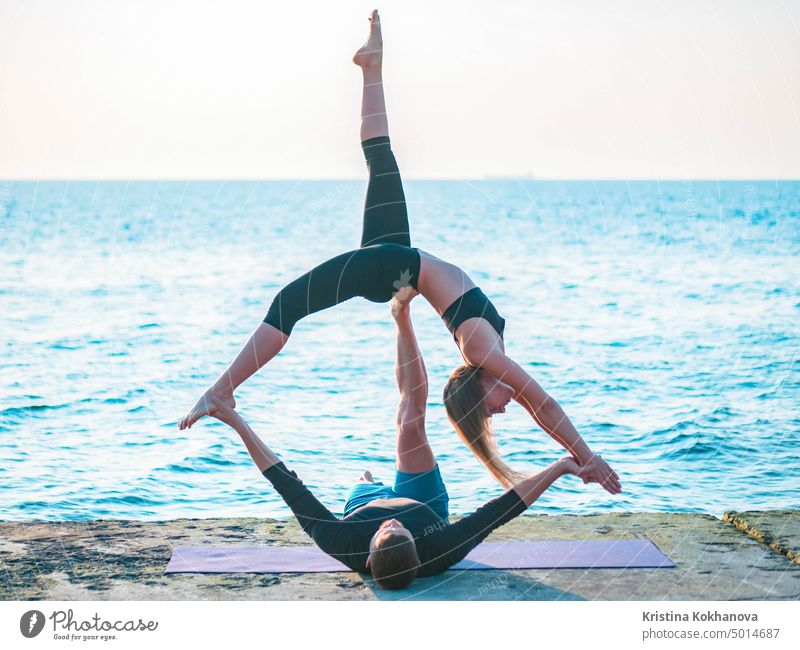 Fit junges Paar macht Acro-Yoga am Meeresstrand. Mann liegt auf Betonplatten und balanciert Frau auf seinen Füßen. Schönes Paar übt zusammen Yoga. acro acroyoga