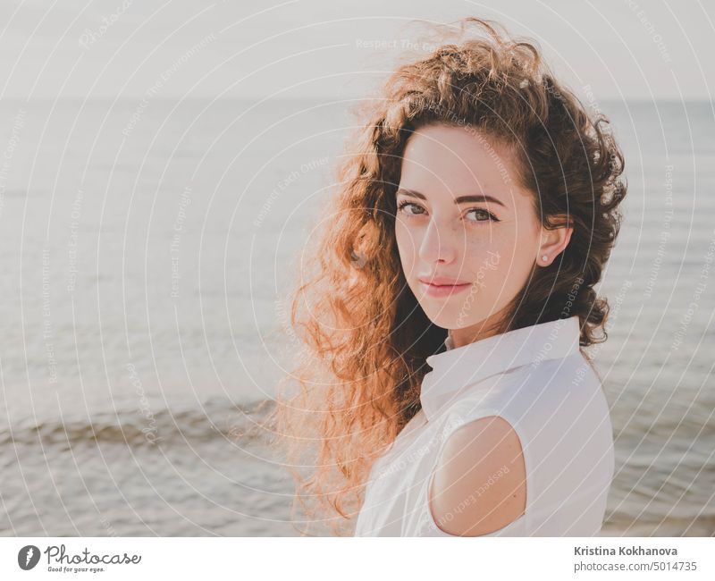 Schönes Mädchen mit lockigem langem Haar in Richtung der Kamera auf sonnigen Frühling Meer Strand. Junge stilvolle Business-Frau in der Mode schwarzen Mantel und weißes Hemd in der Nähe von Meer.