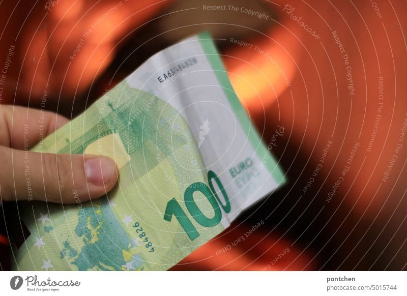 Eine Hand hält einen Geldschein, hundert Euro,  vor einen brennenenden Kamin. Hohe Energiekonzern Energie sparen Bargeld Finanzen heizen kamin verbrennen teuer