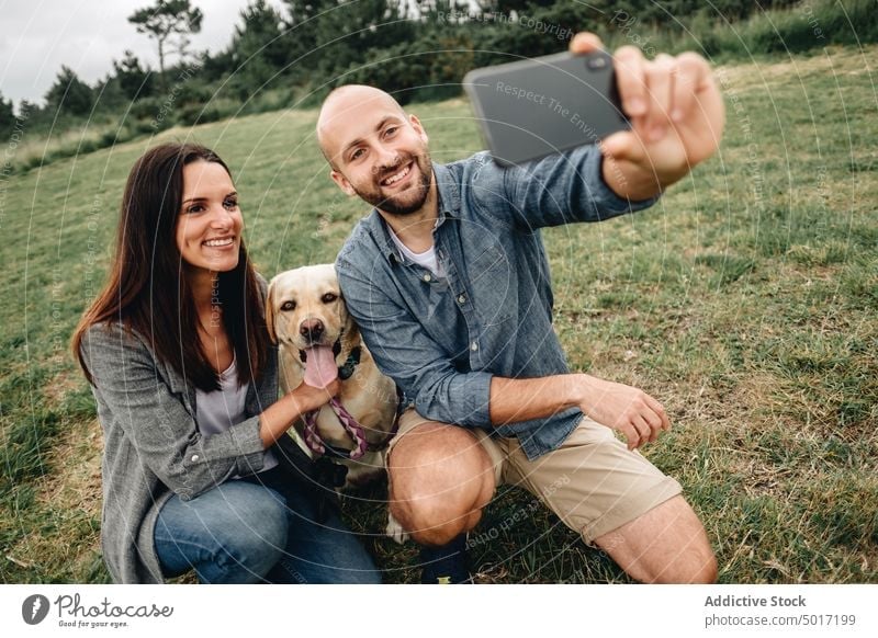 Lächelnde Besitzer sitzen mit Hund und machen ein Selfie im Park Paar Smartphone umarmend Gedächtnis benutzend Haustier Natur grün Gras Tier Labrador Retriever