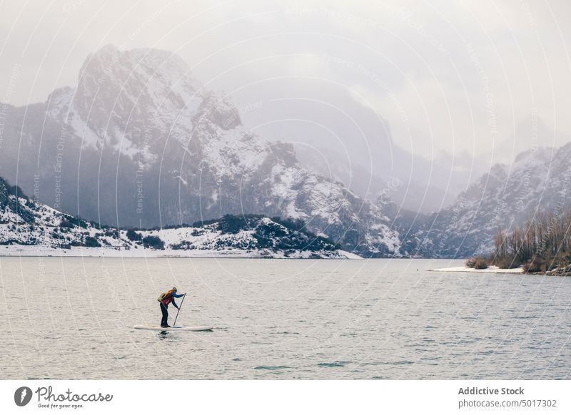Mann auf Paddelbrett zwischen Wasser und Bergen an der Küste Berge u. Gebirge fliegend Tourist Oberfläche Hügel SUP Schnee Ufer Winter malerisch Ansicht Stein