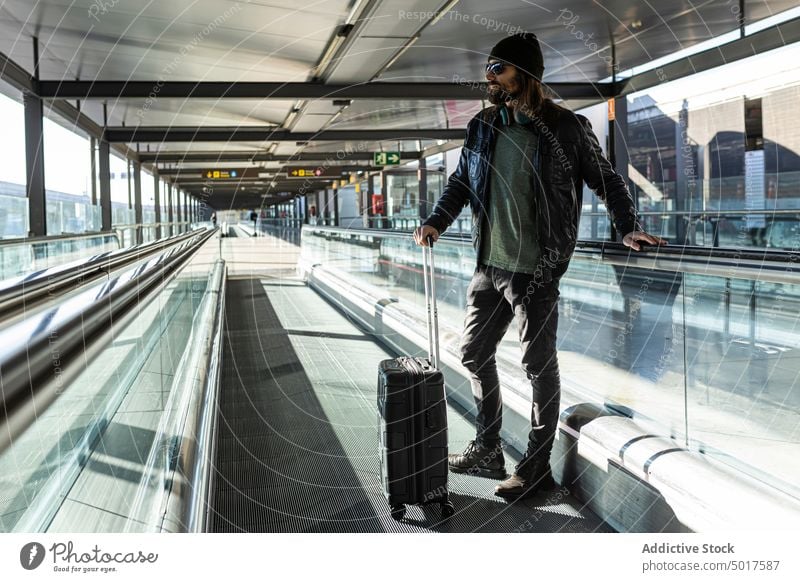 Hipster männlicher Reisender auf dem Laufband im Flughafen Mann reisen Passagier Koffer trendy Stil Erwachsener modern Gepäck Vollbart Hut Lederjacke Ausflug