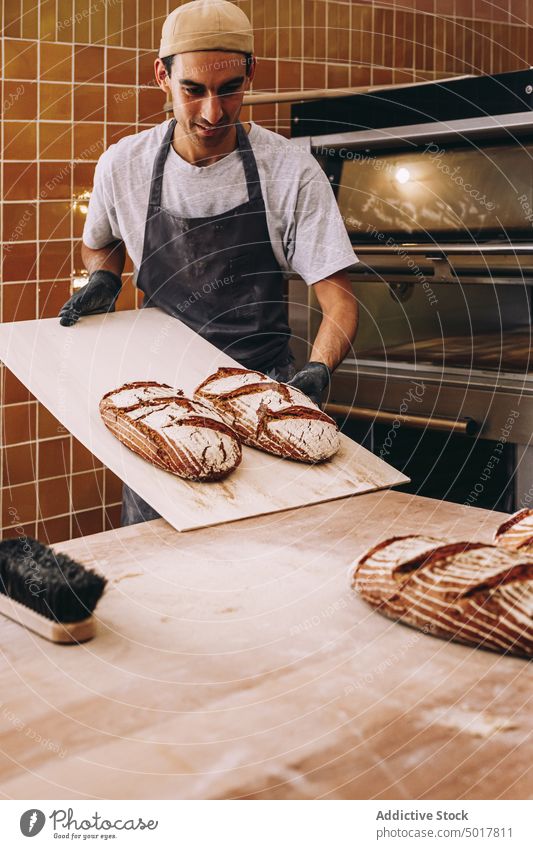 Lächelnder männlicher Koch mit Brot in einer Bäckerei Mann hölzern Tisch gebacken frisch geschmackvoll Ofen heiter Brotlaib Lebensmittel Gebäck lecker Schürze