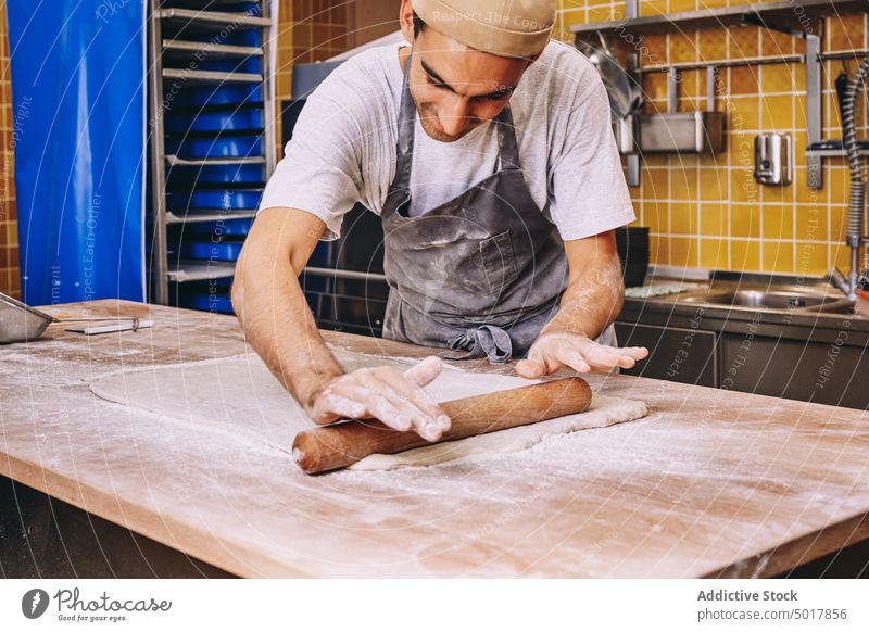 Männlicher Koch, der Teig in einer Bäckerei ausrollt Nudelholz Teigwaren Mann rollen vorbereiten roh Tisch männlich dreckig Mehl Bestandteil Schürze kulinarisch