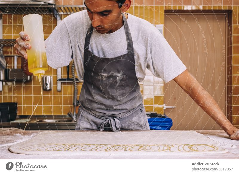 Männlicher Bäcker gießt Öl auf rohen Teig Teigwaren Bäckerei Mann eingießen Erdöl Koch Prozess vorbereiten Rezept männlich Küche Schürze Tisch Arbeit Mehl