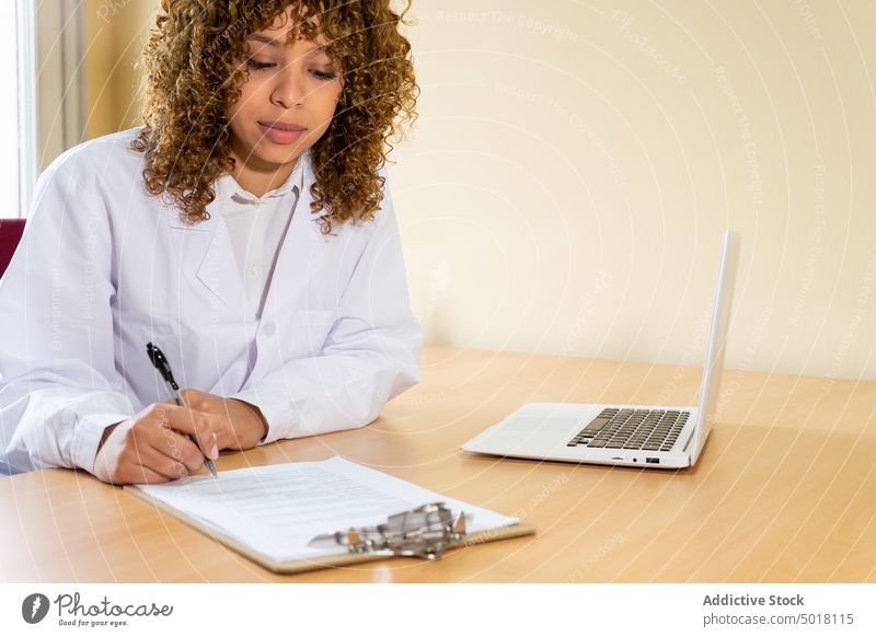 Ethnischer Mediziner schreibt auf Papier am Tisch mit Laptop Sanitäter zur Kenntnis nehmen Schriftstück professionell Arbeit Zwischenablage Frau Büro Klinik