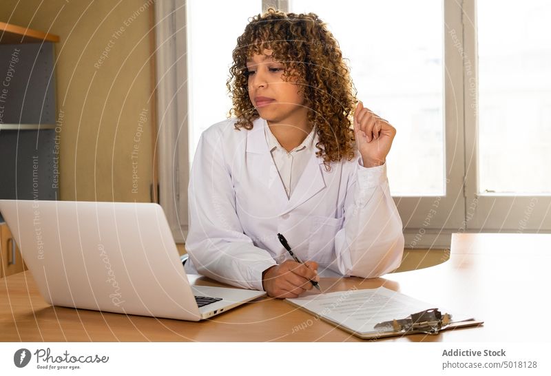 Ethnischer Mediziner schreibt auf Papier am Tisch mit Laptop Sanitäter zur Kenntnis nehmen Schriftstück schreibend professionell Arbeit Zwischenablage Frau Büro